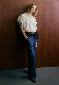 Blonde woman leaning against wooden wall wearing white short-sleeve shirt, blue wide-leg pants, black sweater tied around waist.