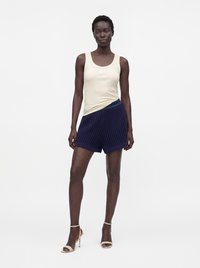Femme debout portant un haut sans manches crème, un short texturé bleu marine et des sandales blanches à talons hauts sur un fond blanc uni.