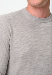 Hellgrauer gestrickter Pullover mit strukturierter Oberfläche, runder Ausschnitt und gerippten Bündchen, der eine Nahaufnahme des Stoffdetails zeigt.