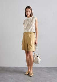 Pletený top bez rukávů v krémové barvě, tanové šortky s vysokým pasem, metalické zlaté mokasíny a malá světle béžová kabelka. Jednoduchý, moderní outfit.