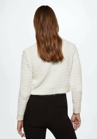 Frau mit langen braunen Haaren, die einen weißen strukturierten Pullover und schwarze Hosen trägt, steht mit dem Rücken zu einem schlichten Hintergrund.