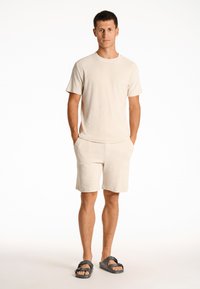 Conjunto de camiseta y pantalones cortos de rizo beige con mangas cortas, corte relajado, bolsillos laterales, combinado con sandalias negras. Textura suave, diseño minimalista.