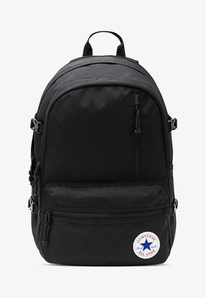 Sac à dos en tissu noir avec plusieurs compartiments zippés, bretelles ajustables et un logo circulaire Converse en bas. Forme rectangulaire basique.