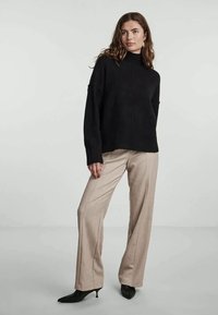 Zwarte gebreide coltrui met een losse pasvorm, gecombineerd met beige wijde broek en zwarte puntige hakken, staand tegen een neutrale achtergrond.