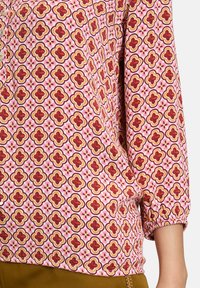 Blouse à manches longues avec un motif floral rose, présentant des formes arrondies en accents rouges et dorés. Les poignets élastiques améliorent l'ajustement.