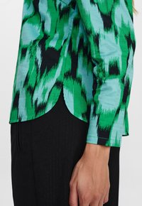 Chemise à manches longues verte et noire avec un ourlet arrondi, fabriquée en tissu léger et à la texture lisse.