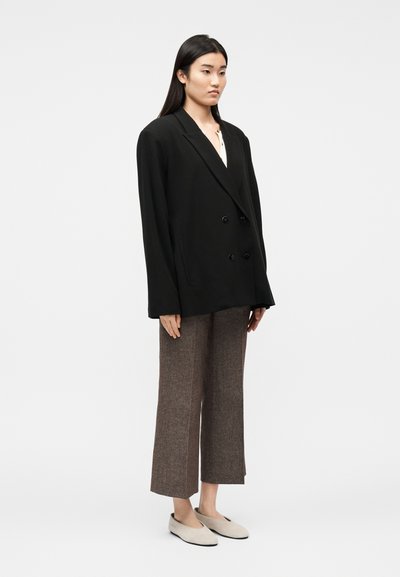 Femme portant un blazer noir oversize à double boutonnage, un pantalon marron à jambes larges et coupé au-dessus de la cheville, et des chaussures plates beiges, debout devant un fond blanc.