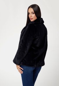 Cappotto in faux fur nera con una consistenza morbida, ampio colletto e vestibilità comoda. Le maniche sono leggermente svasate, abbinato a jeans blu aderenti.