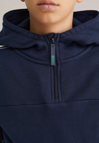 Tumši zils sporta džemperis ar rāvējslēdzēju priekšpusē un teksturētu audumu. Aprīkots ar kapuci un kontrastējošām svītrām uz pleciem.