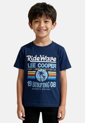 Jeune garçon souriant aux cheveux courts et foncés portant un t-shirt bleu marine avec un motif coloré de surf et de palmier ainsi qu'un texte.