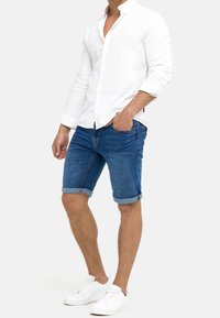 Camicia bianca con bottoni, pantaloni shorts di jeans con orlo arrotolato e sneakers bianche, che mostrano un comodo outfit estivo con una texture di tessuto leggera.
