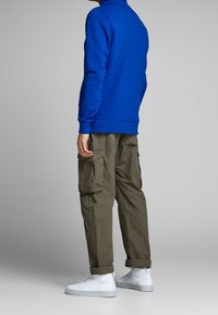 Persona in piedi che indossa un felpa blu, pantaloni cargo verde oliva con risvolti e sneaker alte bianche su uno sfondo semplice.