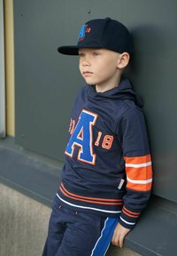 Sinihuppari oransseilla yksityiskohdilla, jossa on suuri chenille-kirjain "A" ja mukana siniset housut. Lapsella on päässään yhteensopiva sininen lippalakki.