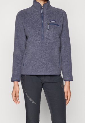 Personne portant un pull polaire bleu Patagonia avec une demi-fermeture éclair et une poche poitrine zippée, associé à un pantalon noir Dynafit.
