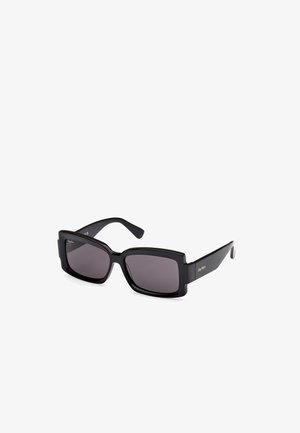 Lunettes de soleil noires carrées avec des verres foncés, monture en plastique épaisse et branding minimal sur la tempête. Texture lisse et design épuré.