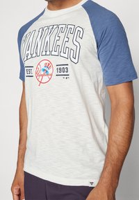 Valkoinen ja sininen lyhythihainen T-paita, jossa on "Yankees"-teksti, logo ja raglanhihat; pehmeä kangas, jossa on heather-kuvio.