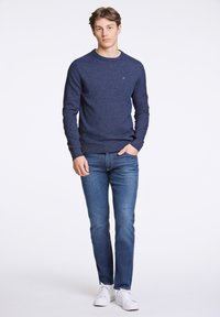 Navy-gestreifter Pullover mit Rundhalsausschnitt, kombiniert mit schmal geschnittenen blauen Jeans und weißen Sneakers, vor einem schlichten Hintergrund stehend.