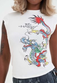 Detailní záběr osoby oblečené v bílém tričku s barevným grafikou draka a textem "Ed Hardy", spárované s tmavými vzorovanými kalhotami.