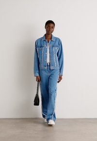 Giacca di jeans blu chiaro con dettagli rovinati, abbinata a pantaloni in denim coordinati con strisce laterali e una borsa a spalla nera.