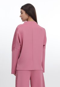 Rosa klær med en glatt tekstur, med blonde detaljer på ermene og høy krage. Designet inkluderer en sømløs bakside.