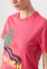 Camiseta de algodón rosa con un diseño gráfico que presenta hojas de palmera verdes, un sol rojo y el texto "PLAYA" en negro y amarillo. Manga corta y cuello redondo.