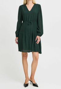 Vihreä, polvipituinen mekko, joka on valmistettu teksturoidusta kankaasta. V-pääntie, pitkät puffihihat, kiristetty vyötärö naruilla ja röyhelöhelma.