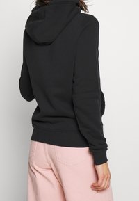 Svart huvtröja med ribbat manschetter och nederkant, tillverkad av mjukt tyg, visad från baksidan med rosa byxor delvis synliga.