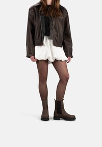 Bruine leren bomberjack, witte gerimpelde shorts, doorschijnende panty’s en bruine enkelaarsjes met elastische zijkanten. Eenvoudig, modern ontwerp.