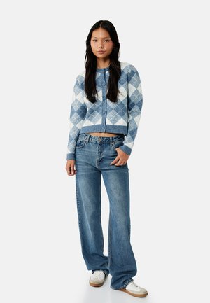 Joven mujer con cárdigan abotonado azul y blanco con rombos, vaqueros de tiro alto y pernera ancha, y zapatillas blancas, de pie sobre fondo blanco.