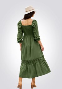 Vestito lungo verde con maniche a sbuffo, corpetto arricciato, bordo in pizzo all'orlo, gonna fluida, abbinato a cappello beige e stivali al ginocchio color cammello.