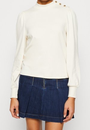 Kobieta ubrana w białą bluzkę z długim rękawem z guzikami na ramionach oraz ciemnoniebieską plisowaną mini spódnicę z dżinsu.