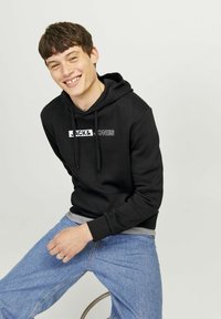 Čierna mikina s kapucňou a predným vreckom, s bielym logom "JACK & JONES". Nosená s svetlomodrými džínami, sediac na stoličke.