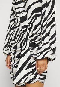 Robe en imprimé zèbre noir et blanc avec des manches amples, un texture froncée à la taille et un détail en nœud. Tissu lisse et léger.