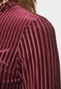 Vinröd topp med vertikala ränder, som har ett texturerat, sammetsliknande tyg. Genomskinliga avsnitt växlar med ogenomskinliga ränder för kontrast.