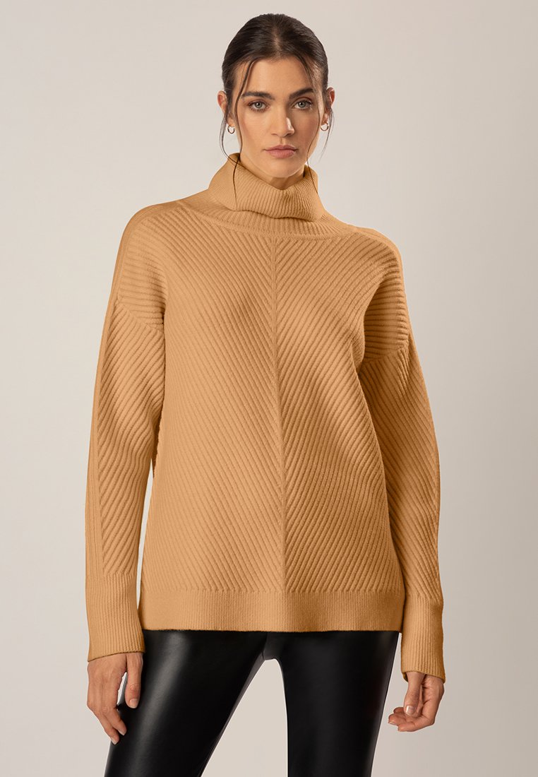 Beige Rollkragenpullover mit diagonaler Rippstruktur, lockerer Passform und langen Ärmeln. Kombiniert mit schwarzen, figurbetonten Hosen.