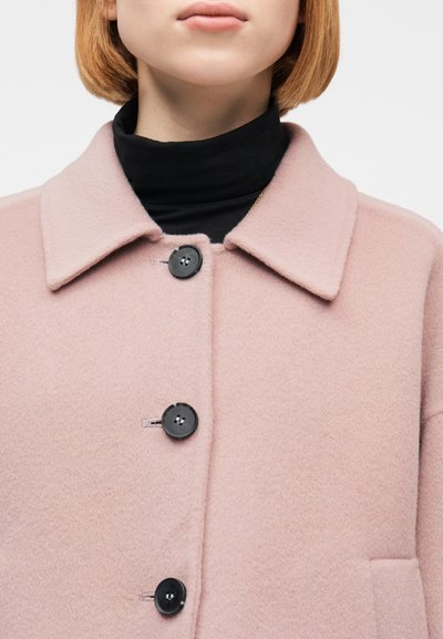 Άτομο που φοράει ανοιχτό ροζ μάλλινο παλτό με μεγάλα μαύρα κουμπιά πάνω από μαύρο πουλόβερ ζιβάγκο, εμφανίζεται από τον λαιμό έως το μέσο του στήθους.