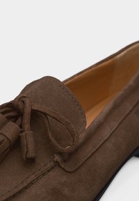 Καφέ δερμάτινο μοκασίνι από σουέντ με διακοσμητικό φιόγκο με φούντες και ταμπά εσωτερική επένδυση, κοντινή άποψη μπροστινού και πλαϊνού μέρους.