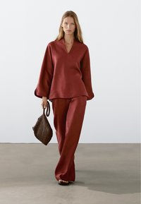 Kobieta idąca w luźnej, ciemnoczerwonej bluzie i spodniach, trzymająca brązową plecioną torebkę, na jednolitym tle i podłodze.