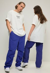Witte T-shirt met korte mouwen met zwarte bedrukte tekst, gecombineerd met losse blauwe broek gemaakt van een gladde stof. Witte sneakers zichtbaar.