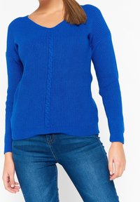 V-ringad stickad tröja i klarblått med strukturerad tyg och en central flätad mönster, tillsammans med blå jeans.