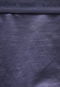 Κοντινό πλάνο σκούρου μπλε βαμβακερού υφάσματος με ζώνη μέσης που φέρει διακριτικό ανάγλυφο κείμενο, εμφανίζοντας πλεκτή υφή και ελαφρείς ρυτίδες.