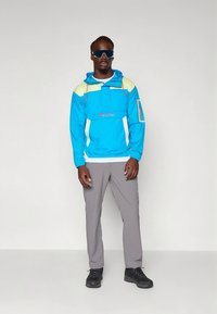 Chubasquero azul y amarillo con un bolsillo frontal y detalles en blanco, combinado con pantalones grises y calzado negro. Gafas de sol completan el conjunto.