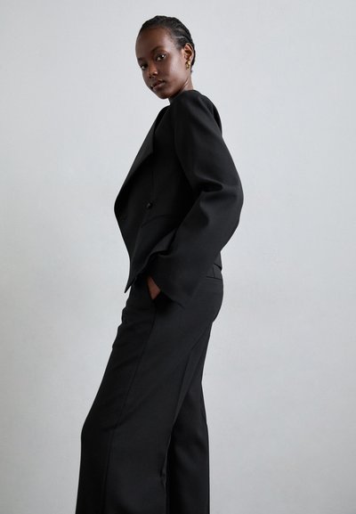 Sort skræddersyet jakkesæt med en tætsiddende blazer med strukturerede skuldre og vide bukser, lavet af glat stof, mod en neutral baggrund.