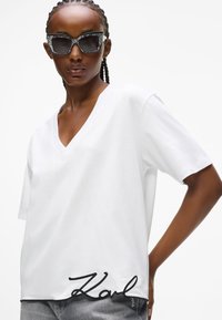 T-shirt bianco a maniche corte con scollatura a V e ricamo nero "Karl" sull'orlo. Abbinato a occhiali da sole neri con motivo.
