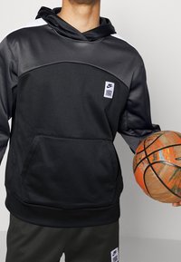 Sweat à capuche noir Nike avec une poche avant, présentant des accents gris et un petit patch logo. Un ballon de basketball tenu, avec un motif orange et vert.