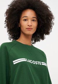 Hanorac verde cu guler rotund și mâneci raglan. Logo-ul alb "LACOSTE" este evidențiat, având două benzi orizontale deasupra.