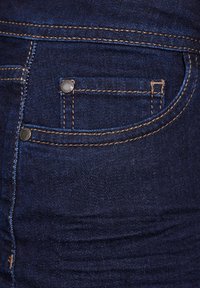 Jeans de mezclilla azul oscuro con una textura suave, que cuentan con un pequeño bolsillo delantero, costuras visibles y detalles de metal.