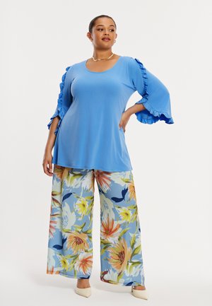 Vrouw staat met hand op heup, draagt een felblauw topje met ruches aan de mouwen en wijde broek met grote oranje en gele bloemen.