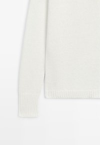 Hvid strikket langærmet sweater med ribstrikkede manchetter og kant, der viser tekstur af stoffet og sy-detaljer.