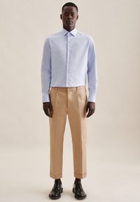 Lys blå langermet skjorte med krage og knappelukking, sammen med beige plisserte bukser og svarte snøresko.